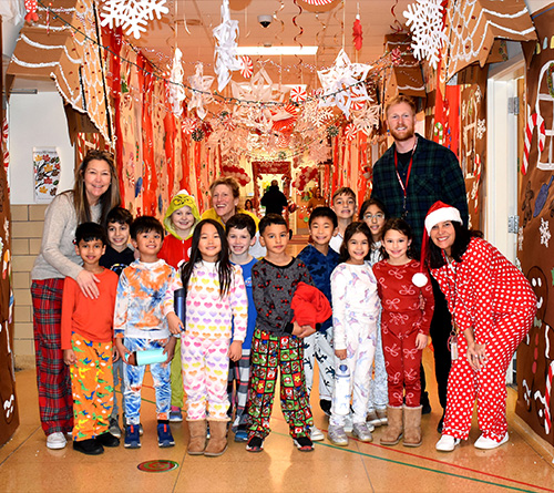 Children and teachers smiling in a festively decorated school hallway with candy-cane and gingerbread holiday displays.