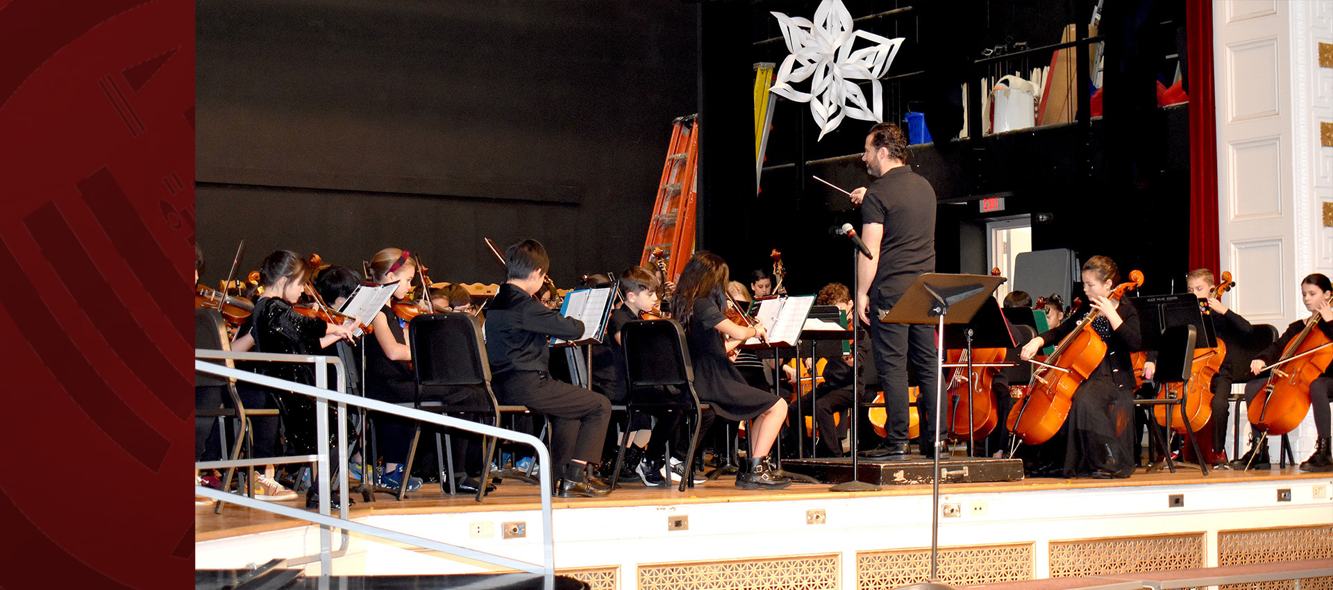 A student orchestra performs on stage with a conductor leading them, as children play violins, cellos, and other string instruments in a concert setting.