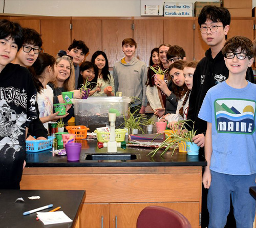 A group of students gather around a classroom table filled with small plants and colorful pots during a hands-on planting activity.