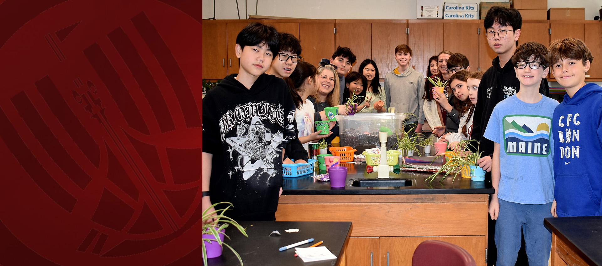 A group of students gather around a classroom table filled with small plants and colorful pots during a hands-on planting activity.