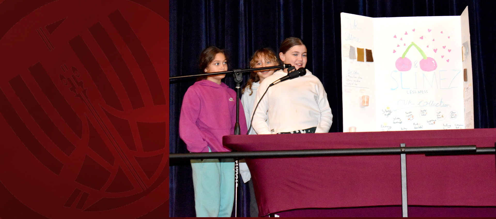 Three children presenting a slime project on stage with a trifold display board.