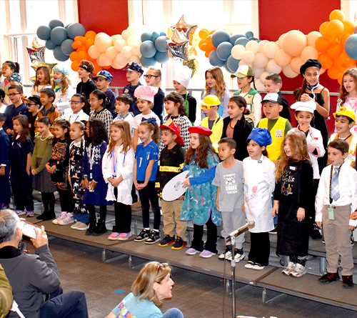 Children dressed in various career costumes stand on risers while families watch and take photos in a decorated event space.