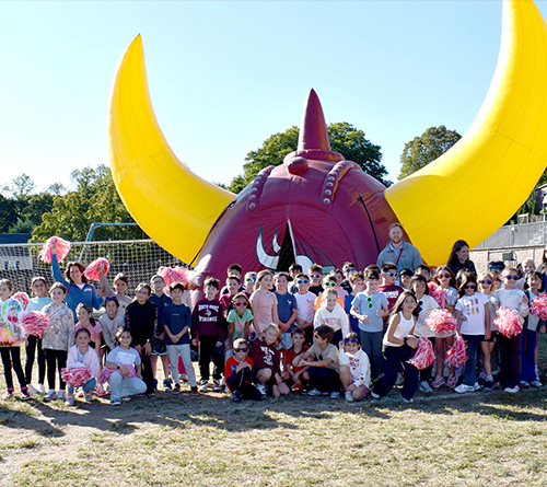 A large group of children and a few adults pose outdoors in front of a giant inflatable Viking helmet with bright yellow horns.