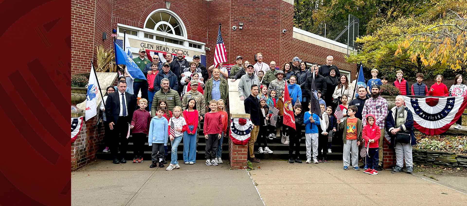A large group of students, families, and veterans gathered on the steps of Glen Head School, holding flags and patriotic banners during a Veterans Day event.