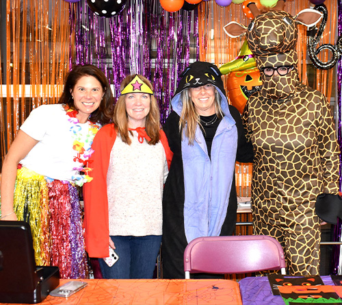 Four adults dressed in colorful Halloween costumes pose together in front of festive decorations with balloons and streamers.