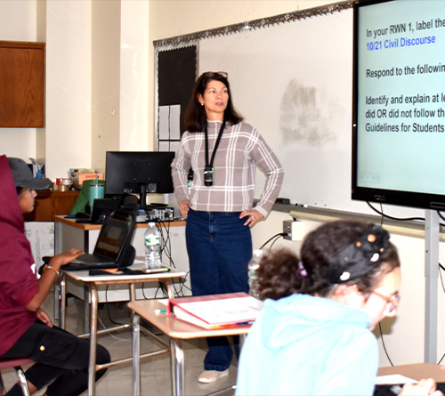 A teacher stands at the front of a classroom beside a large screen displaying an assignment, while students work on laptops at their desks.