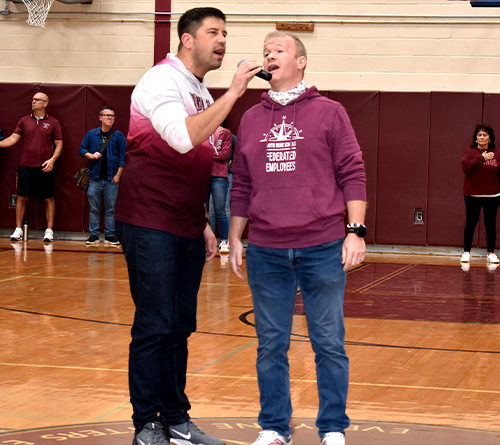 Two men sing into a microphone during a school gym event while others stand in the background.