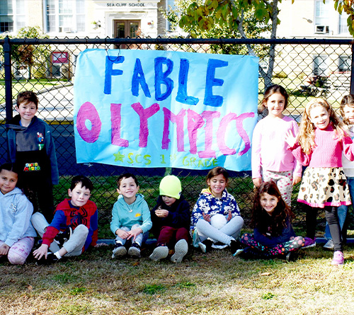A group of young children poses outdoors in front of a handmade banner that reads “Fable Olympics SCS 1st Grade,” with a school building visible in the background.