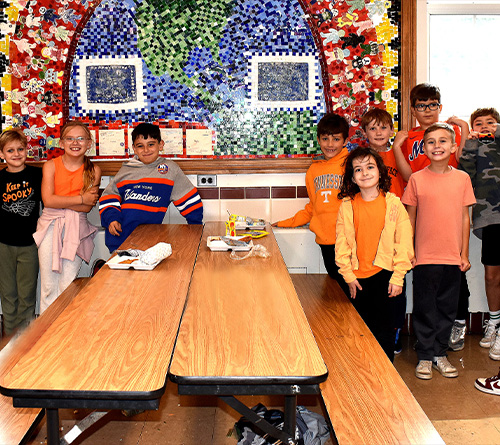 Group of smiling children and a teacher wearing orange clothes posing together in a school cafeteria.