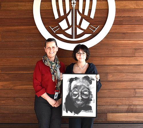 Two people holding a framed artwork in front of a wooden wall
