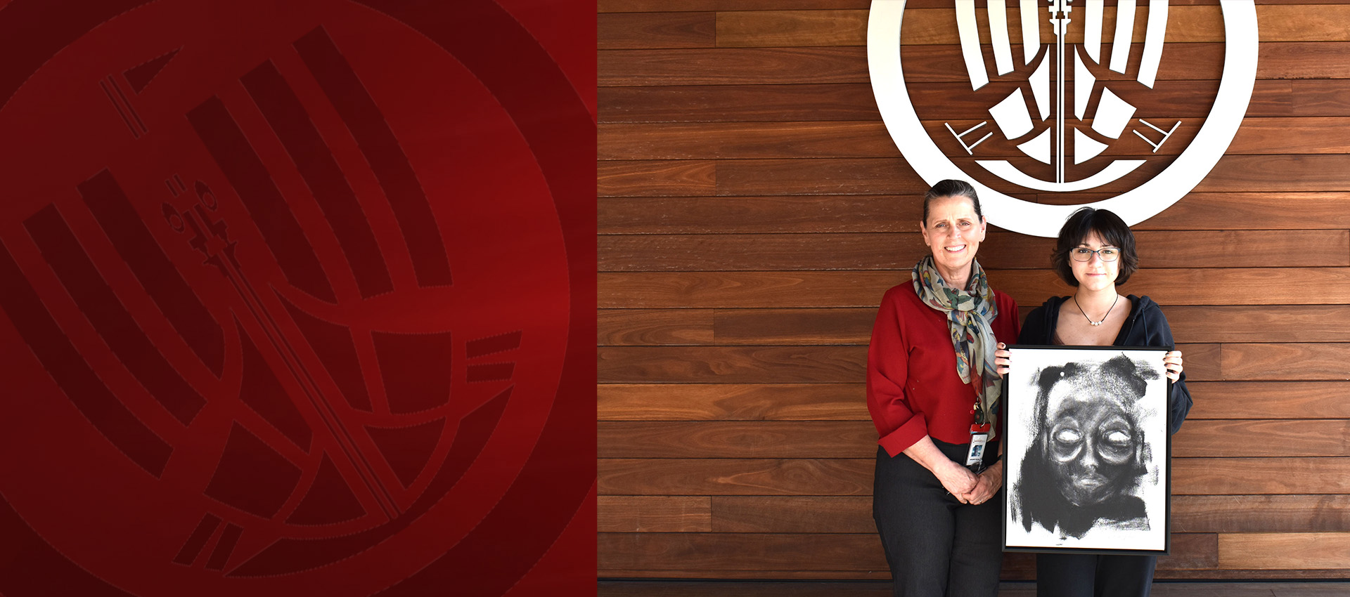 Two people holding a framed artwork in front of a wooden wall