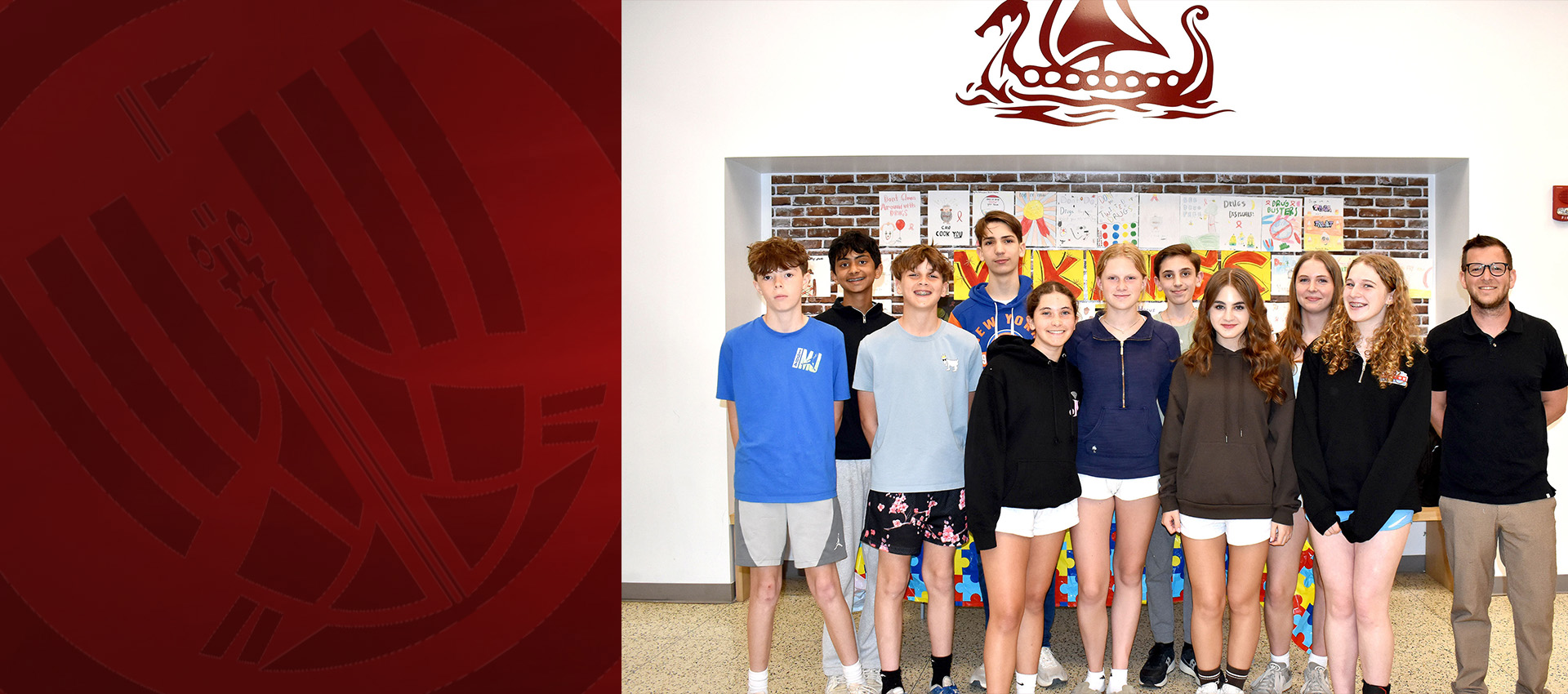 Group of students and a teacher standing together indoors, posing in front of a wall with colorful posters and a Viking ship logo.