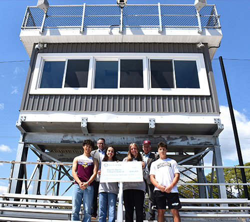 Students and school officials stand in front of a newly constructed high school press box holding a ceremonial grant check at an outdoor athletic field under a clear blue sky.