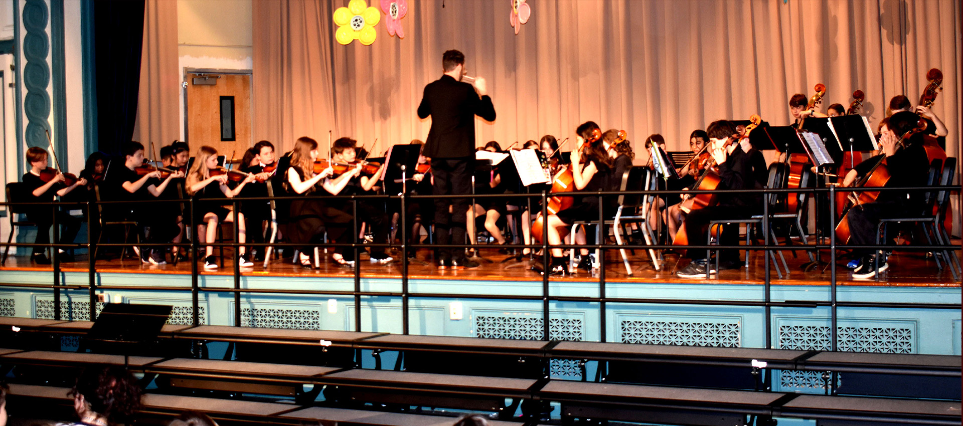 Student orchestra performs on stage during a spring concert, with a conductor leading violin, cello, and bass players dressed in black formal attire.