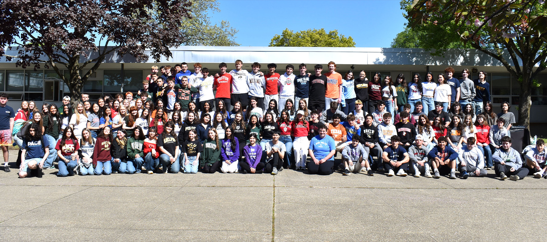 Large group of high school students gathered outside campus wearing college and university shirts.